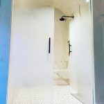 Modern walk-in shower with glass doors featuring decorative film, marble-patterned walls, black fixtures, built-in bench, and white textured floor tiles.
