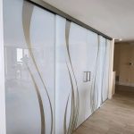 Frosted glass sliding doors with curved beige lines and 3M decorative film separate a hallway from a living area with light wood flooring and neutral walls.