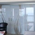 Glass sliding doors enhanced with 3M decorative film feature elegant patterns, separating a modern living area from a balcony that overlooks the ocean; contemporary furniture is visible in the foreground.
