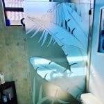 A frosted glass shower door with a tropical leaf design, enhanced by 3M decorative film, stands next to a toilet and shelves, as sunlight streams through a window above.