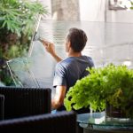 Man using a squeegee on a large glass panel outdoors beside potted plants