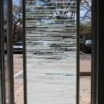 Glass door with horizontal frosted stripes created by 3M decorative film, partially open, revealing parked cars, trees, and a blue sky with clouds outside.