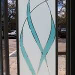 Frosted glass door with turquoise and clear curved lines in an abstract pattern, enhanced by decorative film; parked cars and trees visible outside.
