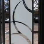 A glass door with frosted white curved patterns made from decorative film, partially clear, stands open to a parking lot with cars and trees visible outside.