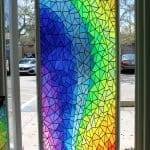 A glass door covered with a mosaic of colorful geometric shapes in a rainbow gradient pattern, sunlight shining through, enhanced by decorative film for a vibrant and modern look.