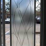 Frosted glass door with intersecting curved line patterns, enhanced by decorative film, partially open to an outdoor parking lot with cars and trees visible in the background.