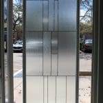 A rectangular glass door with a geometric frosted pattern of 3M decorative film stands open, revealing a parking lot with cars and trees in the background.