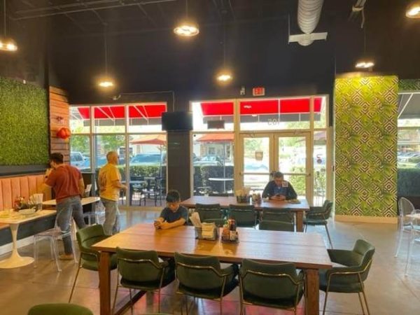 A restaurant interior with people sitting and standing. Tables and chairs are spread out, with some patrons using mobile devices. The room features green and wood accents and large windows with red awnings.