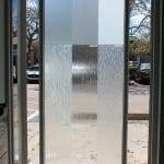 A glass door with six vertical panels, each featuring a unique style of frosted or textured glass, is enhanced with decorative film as sunlight streams in from outside.