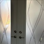 Frosted glass double doors with decorative wavy line patterns created by 3M decorative film and silver door handles, sunlight partially visible through the glass.