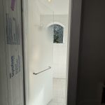 A partially open frosted glass shower door with a metal handle features 3M decorative film in a white-tiled bathroom, sunlight streaming through an arched window.