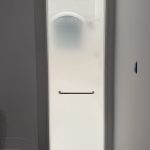 A tall, frosted glass shower door with a metal handle is set in a white bathroom wall; enhanced by 3M decorative film, it allows a faint light to filter softly through the glass.