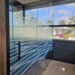 Glass shower enclosure featuring blue and white wave-patterned decorative film, black metal handles, and large windows revealing a tropical outdoor view with palm trees.