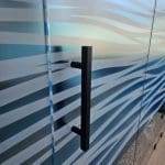 A close-up of a black door handle attached to a glass door covered with 3M decorative film in a blue and white wavy pattern; pebble-patterned flooring is partially visible.