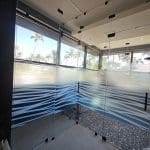 Glass-walled shower with frosted wave patterns created by 3M decorative film, stone floor section, and large windows showcasing palm trees and blue sky outside.