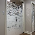 Glass double doors with horizontal frosted stripe patterns created by decorative film, black handle, light wood floor, and closet doors visible in the hallway.