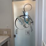 A frosted glass door with an abstract bird design and black swirl pattern, enhanced by decorative film, sits beside a sink and a beige tiled wall in a bathroom.