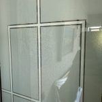 Glass door with a geometric square and rectangle pattern, enhanced by 3M decorative film; faint reflection of a textured wall, picture frame, and indistinct objects visible.