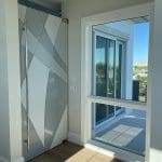 A geometric frosted glass sliding door with 3M decorative film and a large window offering balcony and tree views in a modern, sunlit room.