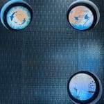 Three round, illuminated porthole displays with aquatic-themed art are mounted on a textured, dark metal wall enhanced by decorative film accents.