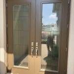 Double brown front doors with geometric glass panels featuring 3M decorative film, metal handles, and a doormat; outdoor scenery and a person reflected in the glass.