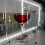 A hand holds a transparent sheet with a printed image of a wine glass filled with red liquid, featuring decorative film, shown in front of a well-lit room with signs and equipment in the background.