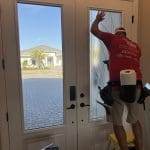 A worker in a red shirt applies decorative film to the glass on a white front door while standing on a yellow step stool inside a house.