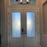 A set of double front doors with frosted glass panels featuring a curved pattern, enhanced by decorative film, beneath a transom window and a black chandelier with white shades.