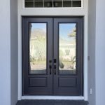 Double front doors with glass panels and black handles, set in a white frame, feature a small rectangular transom window above. Decorative film on the glass adds privacy while still reflecting houses and plants outside.