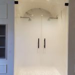 A glass-enclosed shower with double doors, two showerheads, and a white tiled floor features an arched alcove under recessed lighting, enhanced by stylish decorative film for added privacy and flair.