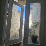 A white front door with 3M decorative film on its frosted glass panels is partially open, revealing a patio with potted plants and a glimpse of blue sky outside.