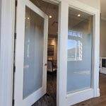 A white-framed glass door with decorative film is open, leading into a carpeted office space with chairs, artwork on the wall, and large windows letting in natural light.