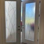 A white front door with frosted glass panels enhanced by a decorative film is partially open, revealing a view of plants and blue sky outside.