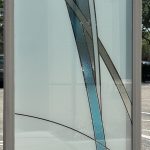 A rectangular frosted glass panel with curved, intersecting lines and blue-tinted sections features decorative film, standing upright outdoors on a sunny day.
