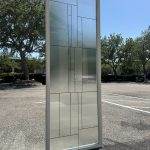 A person holds a rectangular decorative glass window panel with geometric patterns and 3M decorative film in a parking lot on a sunny day.