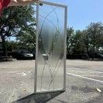 A person holds up a decorative glass panel with an abstract etched design, featuring 3M decorative film, in a parking lot on a sunny day.