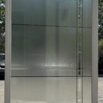 A large rectangular glass door with a white frame is held upright outdoors, reflecting trees, cars, and a basketball hoop. The glass features 3M decorative film for added style and privacy.
