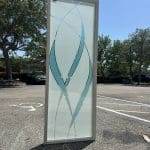 A person holds a decorative glass door panel with blue and clear curved designs, possibly enhanced by 3M decorative film, in a sunny outdoor parking lot.
