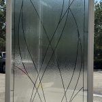 A rectangular glass door panel with a white frame features 3M decorative film in a frosted texture and curved black lines forming an abstract pattern, photographed outdoors.