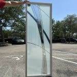 A person holds a tall rectangular glass panel adorned with abstract frosted and clear curved lines created using 3M decorative film, standing in a sunlit parking lot.