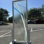 A decorative frosted glass panel with blue and clear geometric lines, enhanced by 3M decorative film, stands on a wooden base in a sunny parking lot.