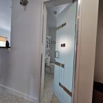 A frosted glass door with 3M decorative film is partly open, revealing a white bathroom with a toilet, cabinet, and tiled floor.