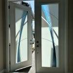 An open white door with frosted glass panels featuring blue abstract designs and 3M decorative film lets in sunlight, revealing an outdoor walkway and paved area.