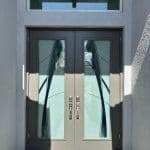 Double gray doors with geometric glass panels enhanced by 3M decorative film, silver handles, and pavers in front, set in a white exterior wall with a transom window above.
