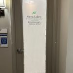 A closed door with frosted glass featuring 3M decorative film labeled “Siena Lakes by Erickson Senior Living, Erickson Health Medical Group,” and a sign nearby reads “Come in, we are open.”.