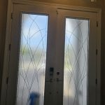 A white double front door with frosted glass panels featuring curved designs, enhanced by decorative film, set below a rectangular transom window with divided panes.