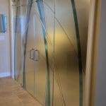 Frosted glass double doors with blue and clear curved line designs, set in a white frame and enhanced by decorative film, separate two indoor spaces with tiled flooring.