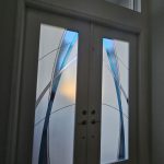 Double doors with frosted glass panels featuring abstract blue and gray curved designs, enhanced by decorative film, set in a white frame with a transom window above.