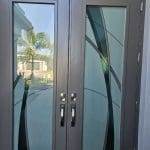 Double front doors with frosted glass panels enhanced by decorative film featuring curved black and green designs, silver handles, and a reflection of a palm tree visible in the glass.