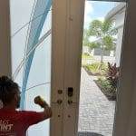 A person is cleaning or applying 3M decorative film to a decorative glass door while seated indoors; the clear glass door offers a view of a sunny yard with palm trees and pavers.